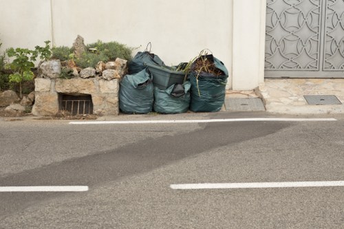 A row of four large, dark green rubbish bags filled with garden waste and debris, positioned on the edge of a paved residential street or driveway next to a white exterior wall. The bags are made of plastic with visible creases and slight tears, and some contain twigs, leaves, and other organic matter. To the left of the bags, there is a small, rectangular garden bed with rocks and plants emerging from the soil, partially enclosed by a low brick or stone border. Behind the bags, the wall features a small grated vent at ground level. On the right, part of a decorative metal gate or door with geometric patterns is visible, indicating an entrance to a property. The scene suggests a garden clearance or rubbish removal activity, with the bags waiting to be collected by Garden Clearance Stockwell, serving the local area near Stockwell, London, SW9.