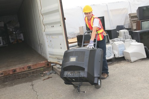 A person wearing a yellow safety helmet, a high-visibility vest with reflective stripes, and gloves is standing outdoors next to a partially open metal storage unit or shipping container. They are holding a portable television with a black plastic casing, a curved screen, and visible controls at the front. The surrounding area includes various discarded electronic items and appliances such as old monitors, computer towers, and miscellaneous debris, suggesting a rubbish removal or clearance operation. The ground is a paved surface with some cracks, and the backdrop features stacked cardboard boxes and building materials. The setting appears to be a commercial or industrial space in Stockwell or nearby London, where Garden Clearance Stockwell is providing rubbish removal services, focusing on clearing electronic waste and other refuse from the site under natural daylight conditions.
