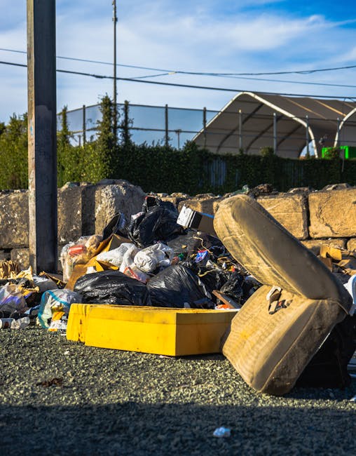 A person wearing a dark beanie, black jacket, and white gloves stands next to a large red metal container filled with black, gray, and orange plastic rubbish bags along with some loose waste materials