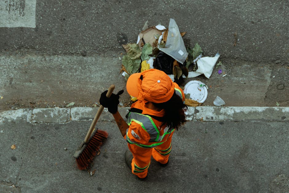 A worker dressed in bright orange high-visibility safety clothing, including a jacket, trousers, and a helmet, is standing on a paved street next to a curb, sweeping litter and debris from the sidewal