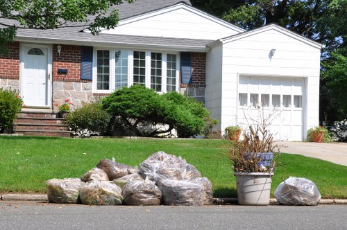 A residential front garden adjacent to a pavement, featuring a neatly maintained lawn with a few small bushes, including a prominent low-lying green shrub near the house. In the foreground, there is a collection of large, irregularly shaped rocks of varying sizes and colors, some with a rough texture, placed along the edge of the pavement. To the right of the rocks, a white plastic bucket contains dried branches or garden debris. The house in the background is a single-story structure with white siding and a brick facade around the windows and front door, which is accessed via a small brick staircase. The property includes a driveway leading up to a white garage door, with potted plants arranged near the base of the garage. The overall scene is well-lit, indicating a sunny day, and the image visually supports services related to rubbish or garden clearance by Garden Clearance Stockwell, located in or near London. The setting appears to be in an urban or suburban part of Stockwell, with typical residential features and a tidy appearance.