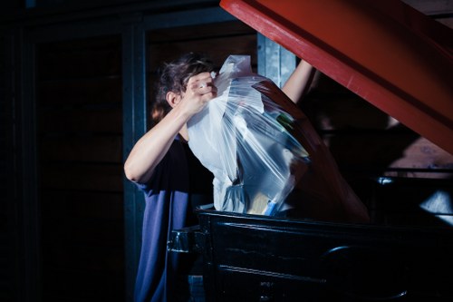 A person wearing a dark shirt is placing a large transparent plastic bag filled with various waste materials into a black mobile rubbish bin, with the bin's lid open. The environment appears to be a storage or garage area with dark wooden and metal surfaces in the background, indicating a clean and organized space used for rubbish collection. The plastic bag contains mixed waste, with some visible papers or packaging, and the person is partially visible, focusing on the action of disposing of rubbish. The scene reflects the process of rubbish removal, consistent with services offered by Garden Clearance Stockwell, particularly for domestic or garden waste clearance in the Stockwell area.