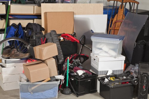 A collection of various household items and storage boxes arranged together in a cluttered indoor space, likely for rubbish removal purposes. The scene features cardboard boxes, plastic storage containers, and black toolboxes, some stacked or placed side by side, with a bicycle wheel partially visible on the left. Several items appear to be packed or ready for disposal, including what looks like fabric, paper, and plastic wrapped materials. In the background, there are shelves with books, and a wooden chair partially visible on the right. The environment suggests a garage or storage area, with a mix of textures including cardboard, plastic, metal, and fabric, illuminated by natural or overhead light, reflecting a typical setting where Garden Clearance Stockwell might carry out rubbish removal services in London or surrounding areas.